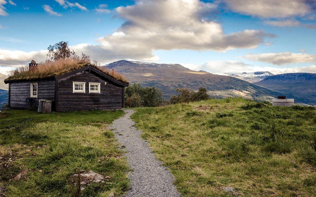 En idyllisk, mørkbeiset hytte med torvtak i et vakkert fjell-landskap, brukt til Spira Takst sin sommerhilsen.